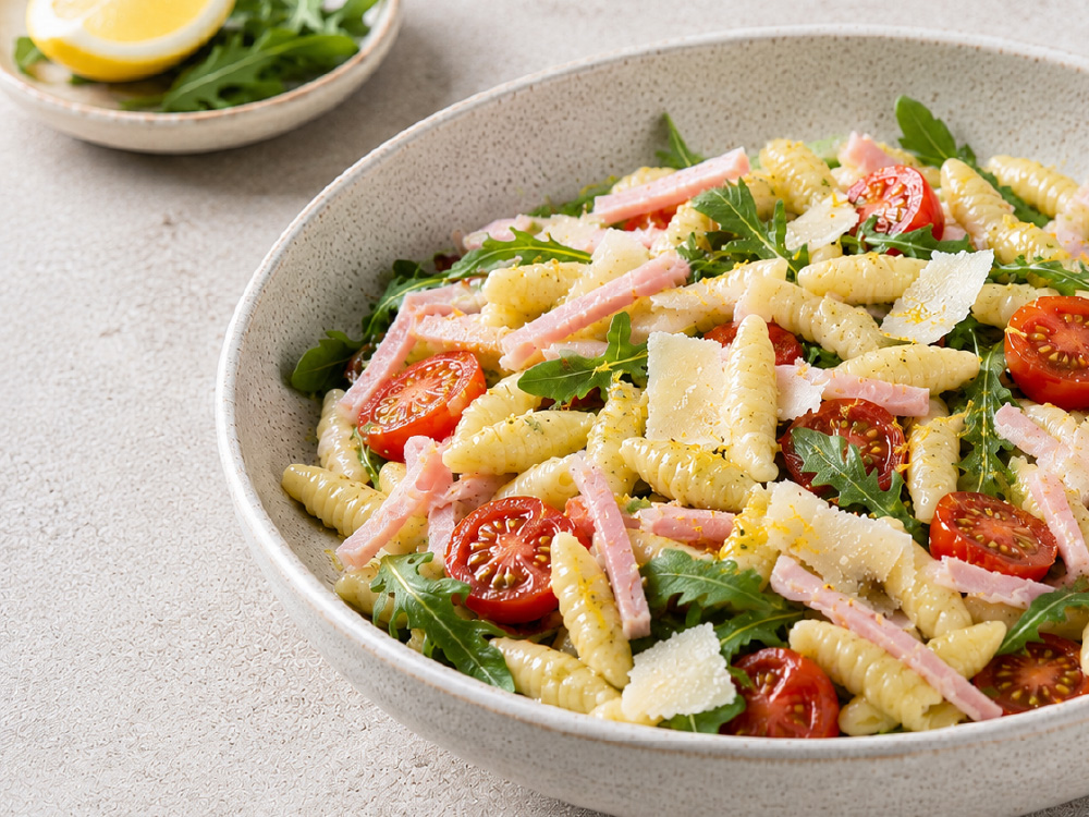 Un piatto abbondante di pasta con prosciutto cotto, pomodorini, rucola e scaglie di parmigiano.